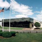 Front view of a modern office building with a reflective glass facade, two flagpoles (American flag and another flag) and a landscaped lawn.