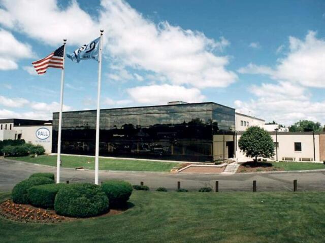 Front view of a modern office building with a reflective glass facade, two flagpoles (American flag and another flag) and a landscaped lawn.