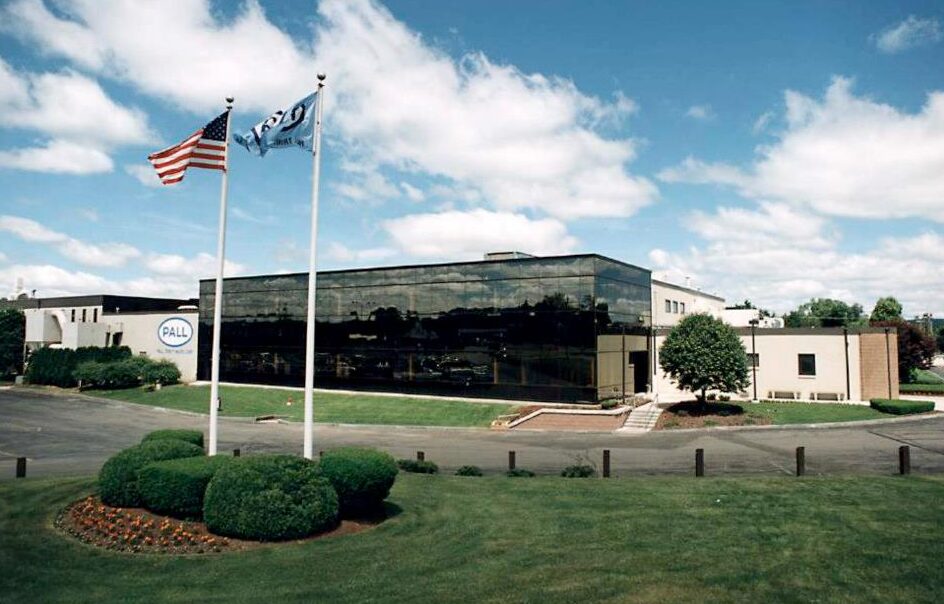 Front view of a modern office building with a reflective glass facade, two flagpoles (American flag and another flag) and a landscaped lawn.