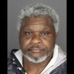 Mugshot photo of an African American man with gray hair and beard, wearing a dark jacket over a light hoodie, neutral expression against a gray backdrop.