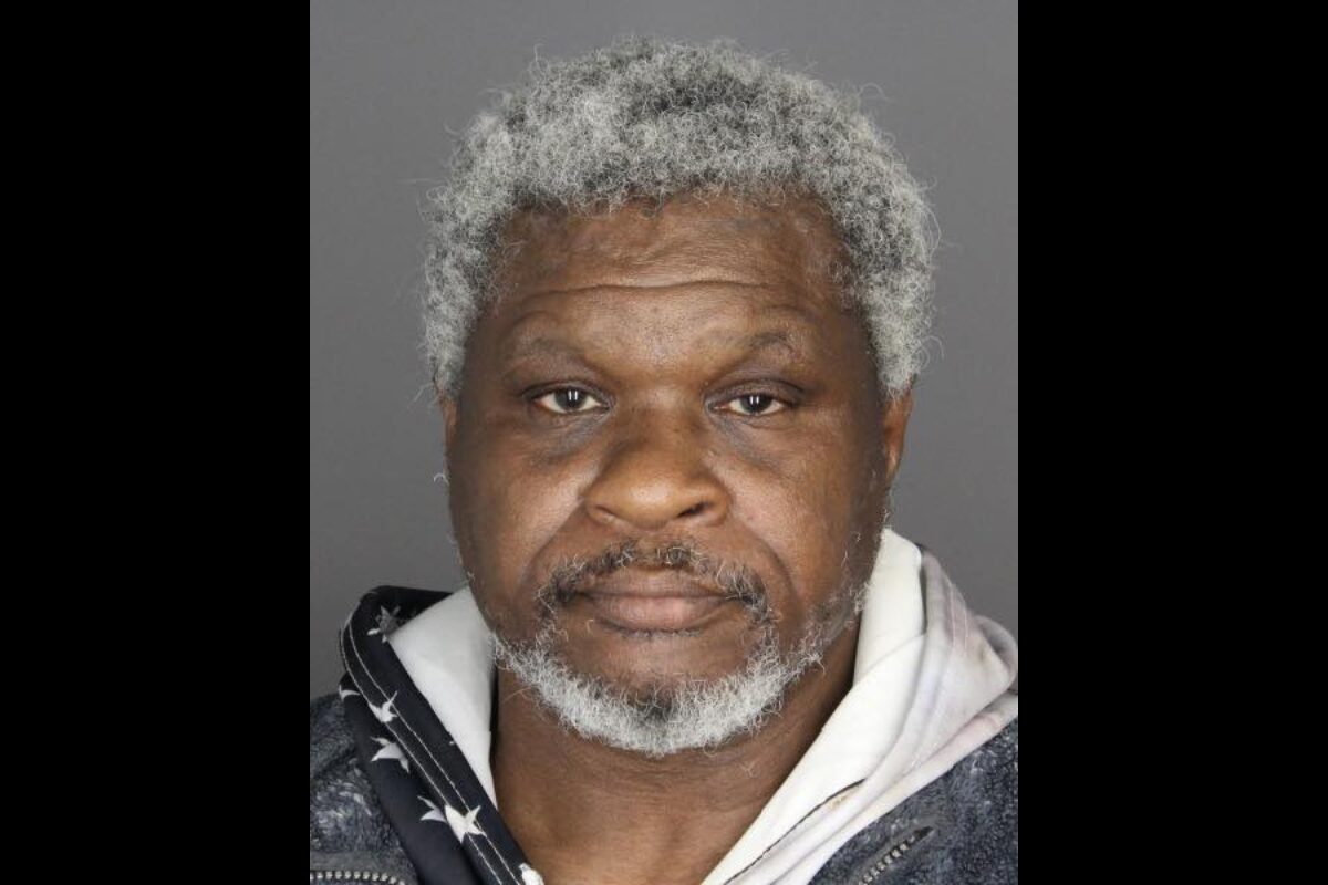 Mugshot photo of an African American man with gray hair and beard, wearing a dark jacket over a light hoodie, neutral expression against a gray backdrop.