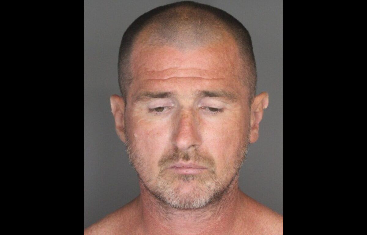 Mugshot of a middle-aged man with a shaved head and light stubble, looking downward against a gray background.