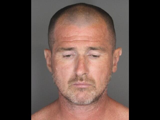 Mugshot of a middle-aged man with a shaved head and light stubble, looking downward against a gray background.