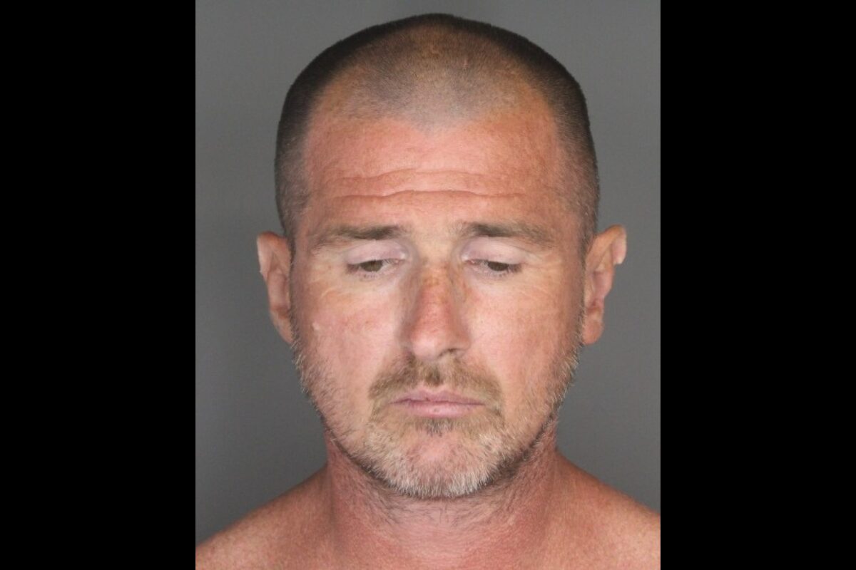 Mugshot of a middle-aged man with a shaved head and light stubble, looking downward against a gray background.