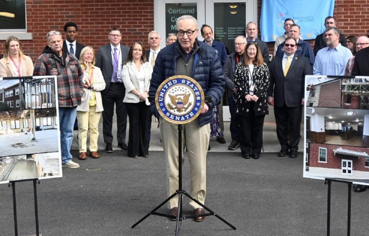 Senator speaking at a podium with the U.S. Senate seal, flanked by a group of officials outside a brick building, with construction posters on either side.