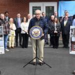 Senator speaking at a podium with the U.S. Senate seal, flanked by a group of officials outside a brick building, with construction posters on either side.
