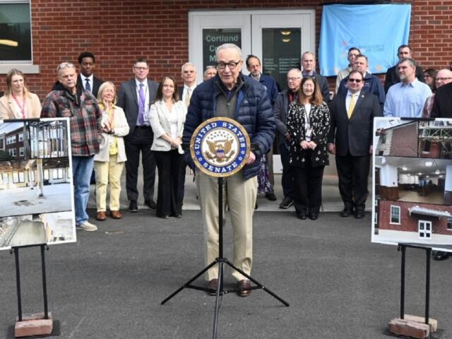 Senator speaking at a podium with the U.S. Senate seal, flanked by a group of officials outside a brick building, with construction posters on either side.