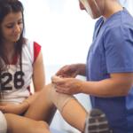 Healthcare professional in blue scrubs bandages a knee for a seated female athlete with a volleyball nearby.