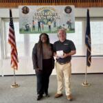 Two smiling adults stand indoors in front of a 'Earth Month' banner with American and state flags behind them.