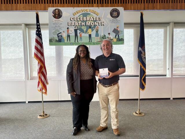 Two smiling adults stand indoors in front of a 'Earth Month' banner with American and state flags behind them.
