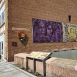 Brick visitor center with a large mural on the side wall, NPS shield, and a row of benches along a paved walkway.