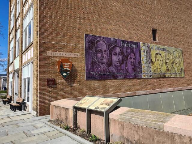 Brick visitor center with a large mural on the side wall, NPS shield, and a row of benches along a paved walkway.