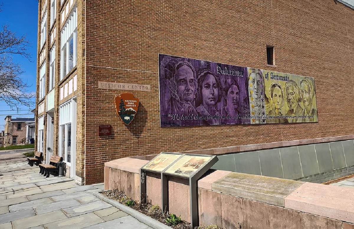 Brick visitor center with a large mural on the side wall, NPS shield, and a row of benches along a paved walkway.