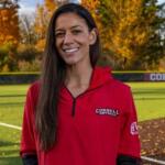 Smiling woman in a red Cornell softball pullover standing on a baseball field with autumn trees in the background, a sunny day.