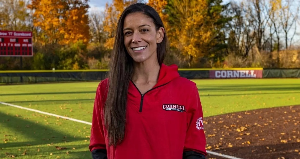 Smiling woman in a red Cornell softball pullover standing on a baseball field with autumn trees in the background, a sunny day.