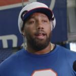 Male athlete being interviewed indoors, wearing a blue shirt and a towel wrapped on his head, speaking to a Bills microphone-mast nearby.