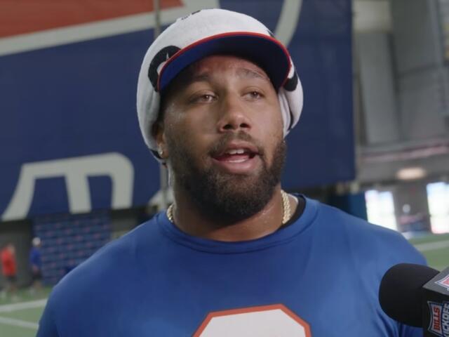 Male athlete being interviewed indoors, wearing a blue shirt and a towel wrapped on his head, speaking to a Bills microphone-mast nearby.