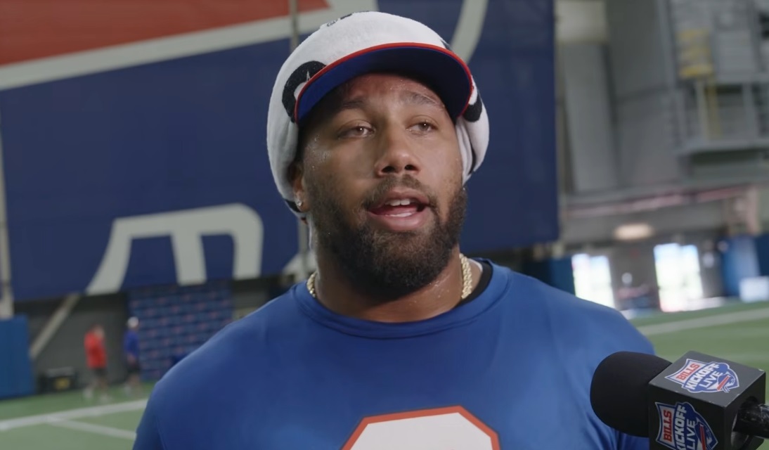 Male athlete being interviewed indoors, wearing a blue shirt and a towel wrapped on his head, speaking to a Bills microphone-mast nearby.