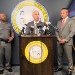 Officials hold a press conference at a podium with microphones, flanked by the county seal and flags behind them, in a formal room.