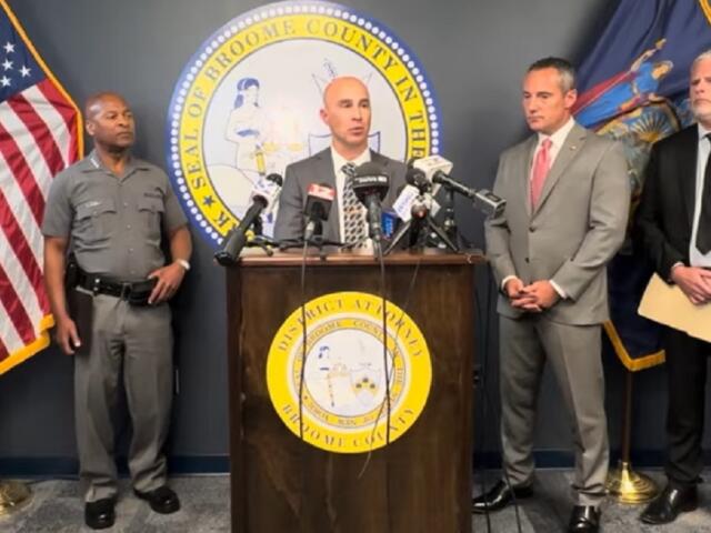 Officials hold a press conference at a podium with microphones, flanked by the county seal and flags behind them, in a formal room.