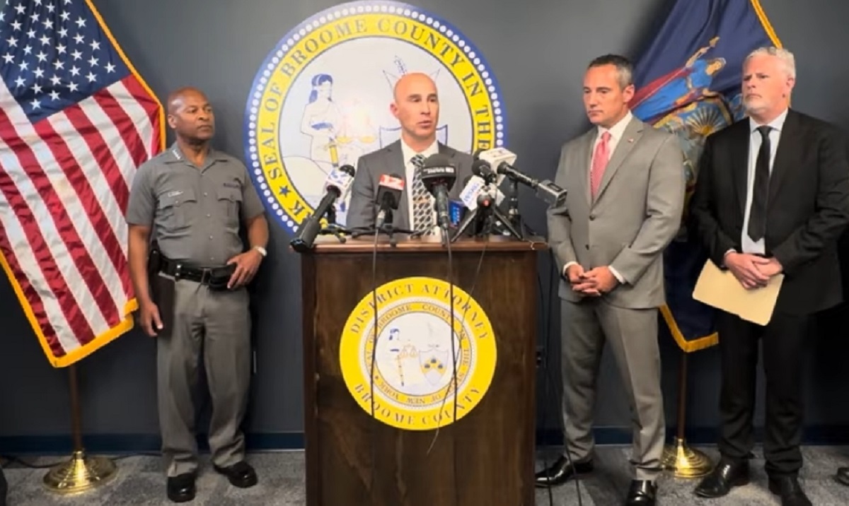 Officials hold a press conference at a podium with microphones, flanked by the county seal and flags behind them, in a formal room.