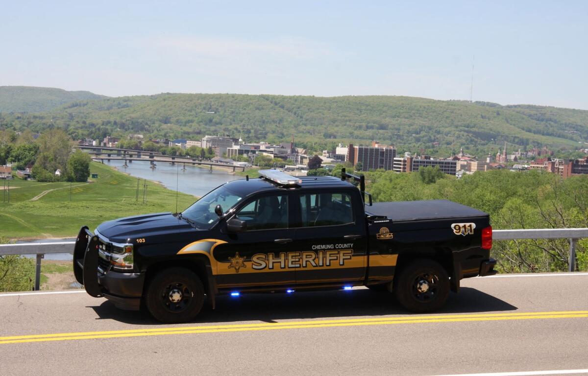 Chemung County Sheriff pickup truck parked on a road, with a river, city buildings, and forested hills in the background.