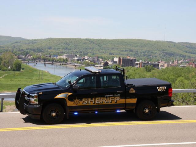 Chemung County Sheriff pickup truck parked on a road, with a river, city buildings, and forested hills in the background.