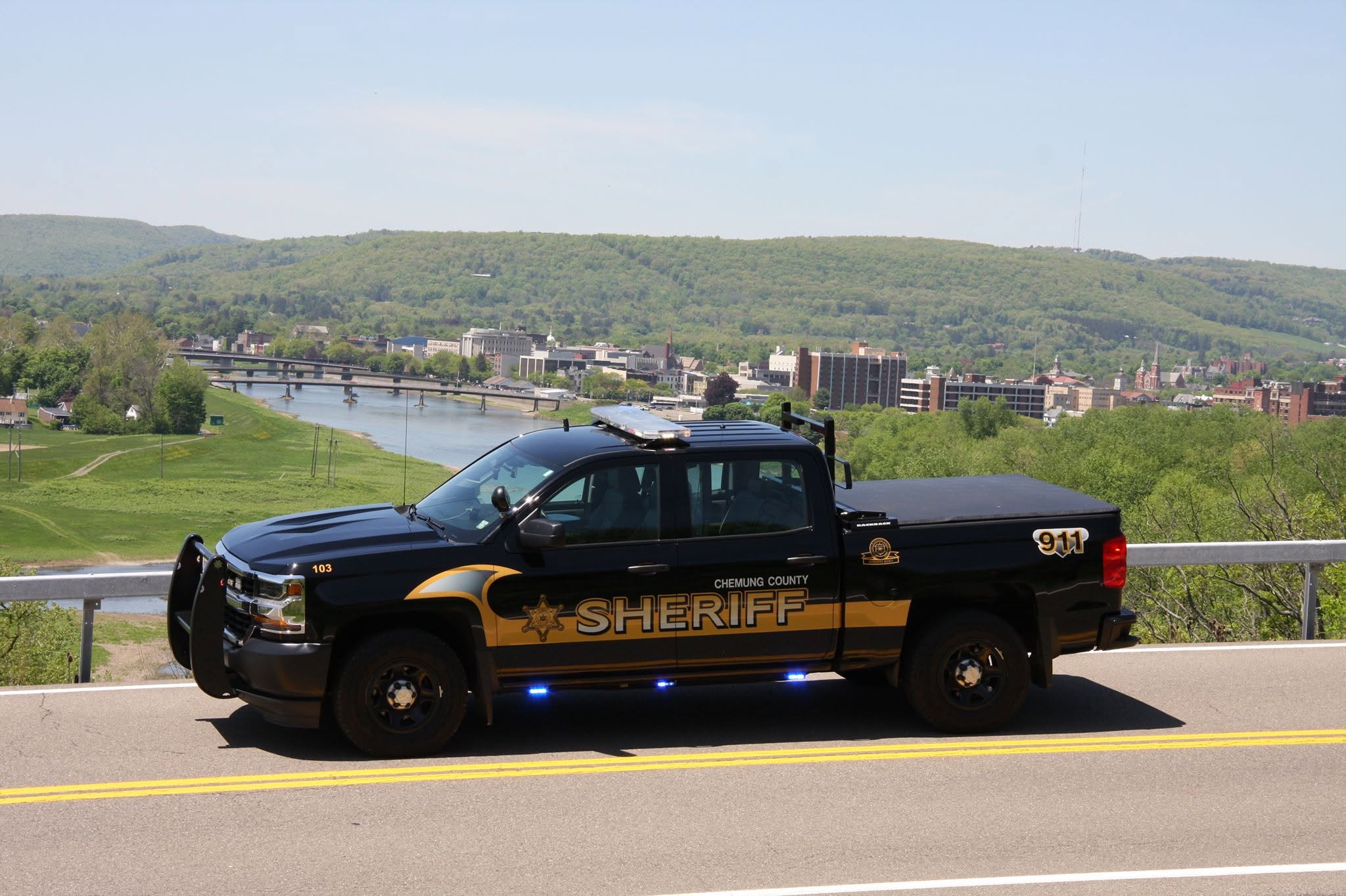 Chemung County Sheriff pickup truck parked on a road, with a river, city buildings, and forested hills in the background.