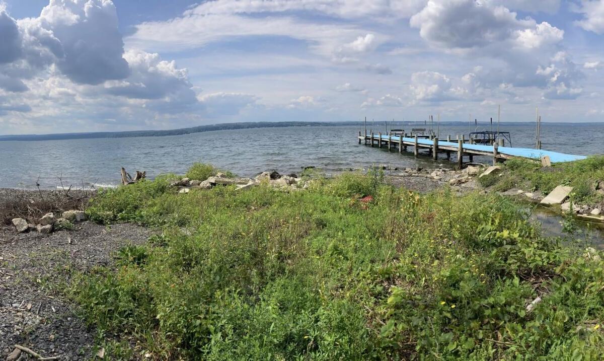 Panoramic view of a rocky beach along a lake with a wooden dock extending into the water and cloudy blue sky above.