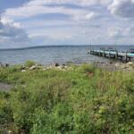 Panoramic view of a rocky beach along a lake with a wooden dock extending into the water and cloudy blue sky above.