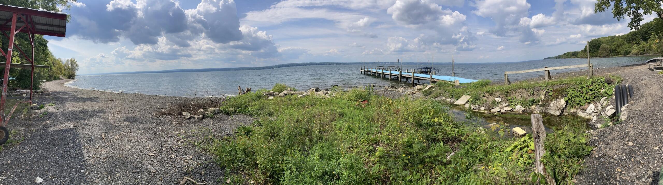 Panoramic view of a rocky beach along a lake with a wooden dock extending into the water and cloudy blue sky above.