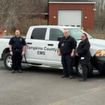Three emergency medical technicians stand beside a white Tompkins County EMS pickup truck in a parking lot.