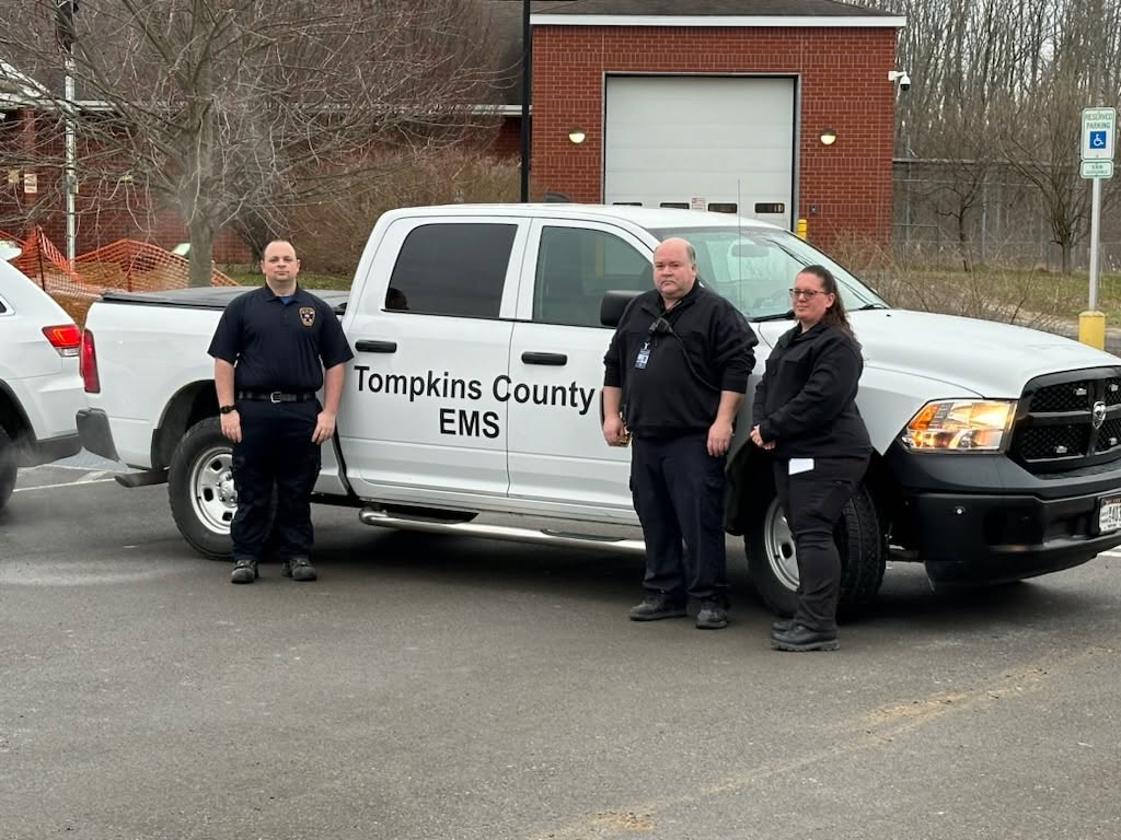 Three emergency medical technicians stand beside a white Tompkins County EMS pickup truck in a parking lot.