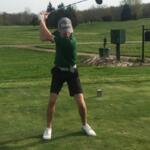 Golfer in a green shirt and cap in the middle of a backswing on a tee box, with a bag and golf course in the background.