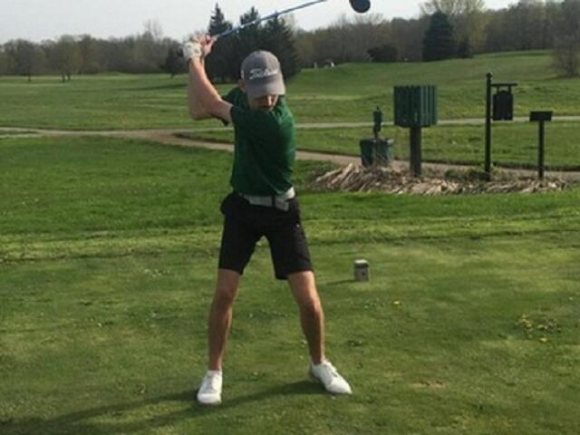 Golfer in a green shirt and cap in the middle of a backswing on a tee box, with a bag and golf course in the background.