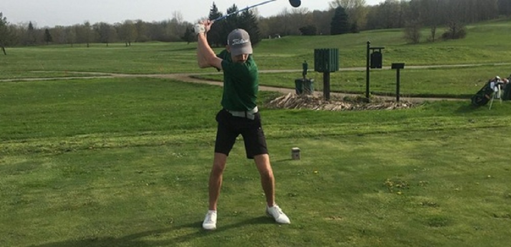 Golfer in a green shirt and cap in the middle of a backswing on a tee box, with a bag and golf course in the background.