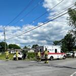 Emergency responders and a white ambulance on a street, with firefighters and fire trucks at the scene under power lines.