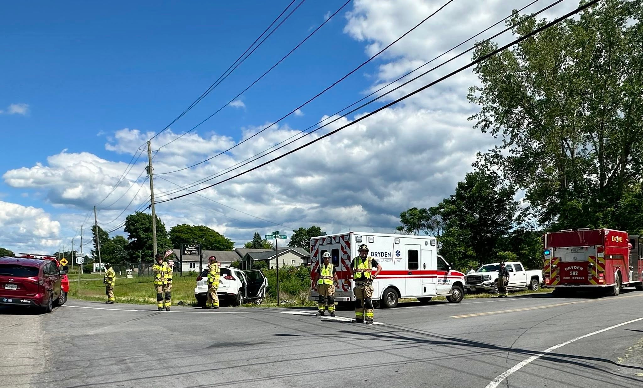 Emergency responders and a white ambulance on a street, with firefighters and fire trucks at the scene under power lines.