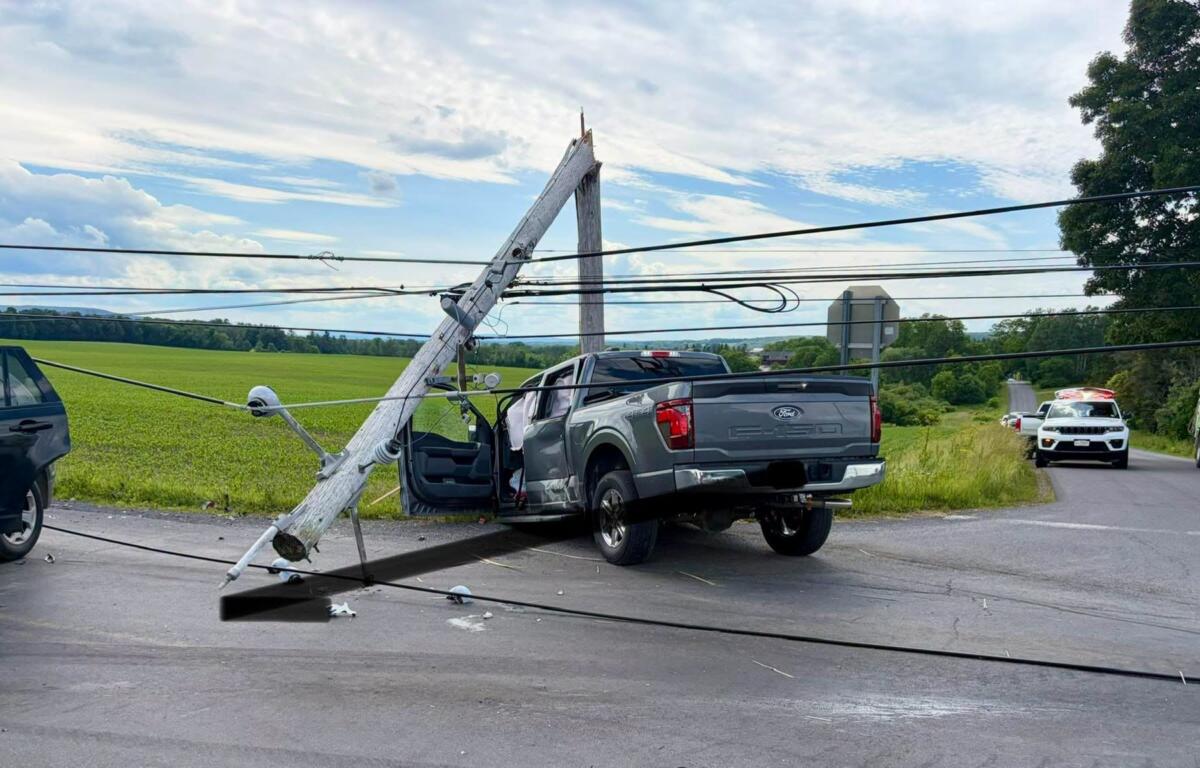 Leaning wooden utility pole fallen onto a gray Ford F-150 pickup on a rural road; power lines tangled, with emergency vehicles in the distance nearby.