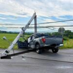 Leaning wooden utility pole fallen onto a gray Ford F-150 pickup on a rural road; power lines tangled, with emergency vehicles in the distance nearby.
