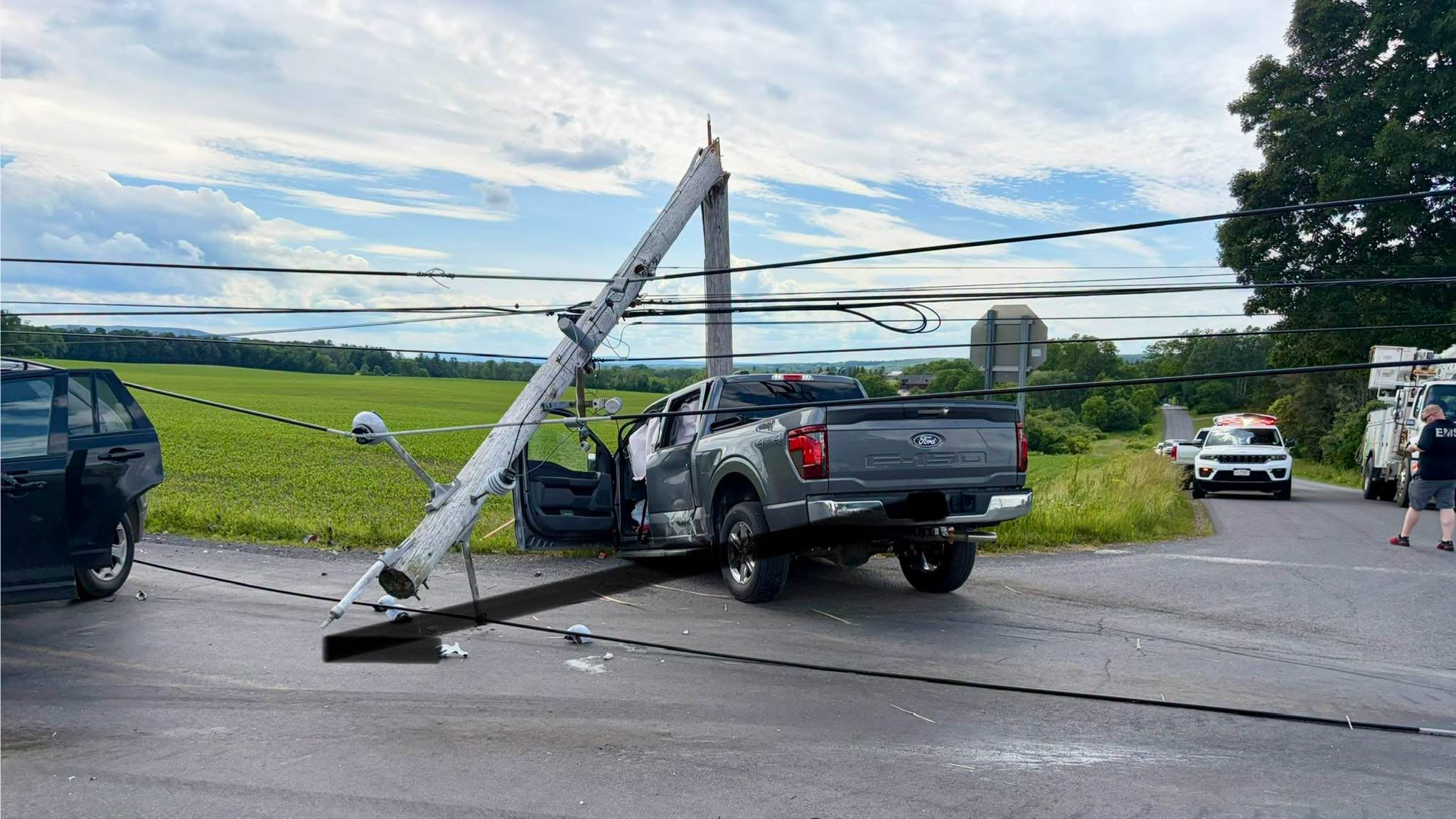 Leaning wooden utility pole fallen onto a gray Ford F-150 pickup on a rural road; power lines tangled, with emergency vehicles in the distance nearby.