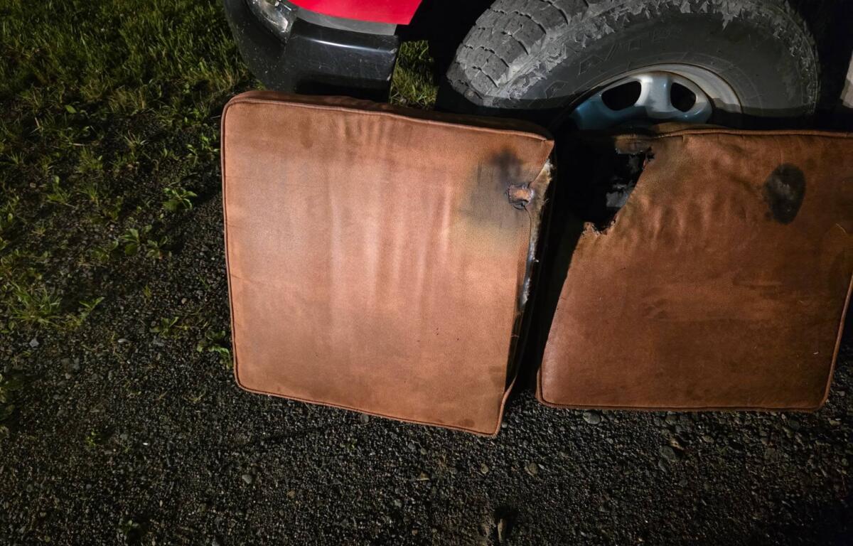 Red vehicle bumper visible with a large treaded tire resting beside brown cushions on a gravel surface. Grass is on the left.