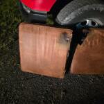 Red vehicle bumper visible with a large treaded tire resting beside brown cushions on a gravel surface. Grass is on the left.