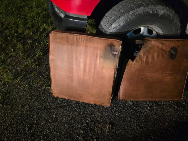 Red vehicle bumper visible with a large treaded tire resting beside brown cushions on a gravel surface. Grass is on the left.