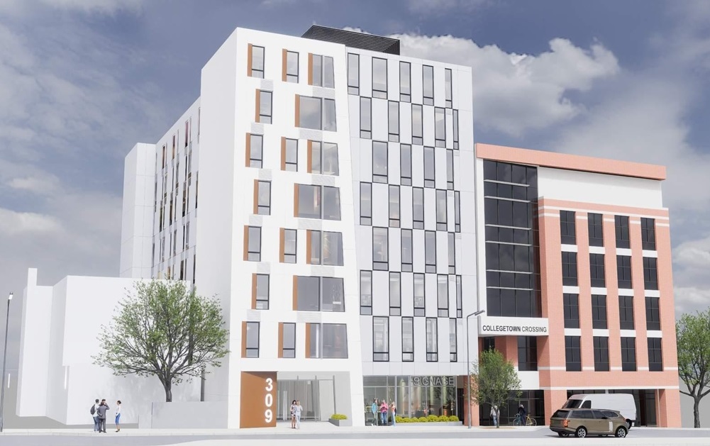 Contemporary mixed-use building with white and brick facades, tall windows, and pedestrians on the sidewalk under a blue sky; Collegetown Crossing sign visible.