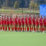 Girls' soccer team in red uniforms lined up on a field, with two neon yellow goalkeepers on the right side.
