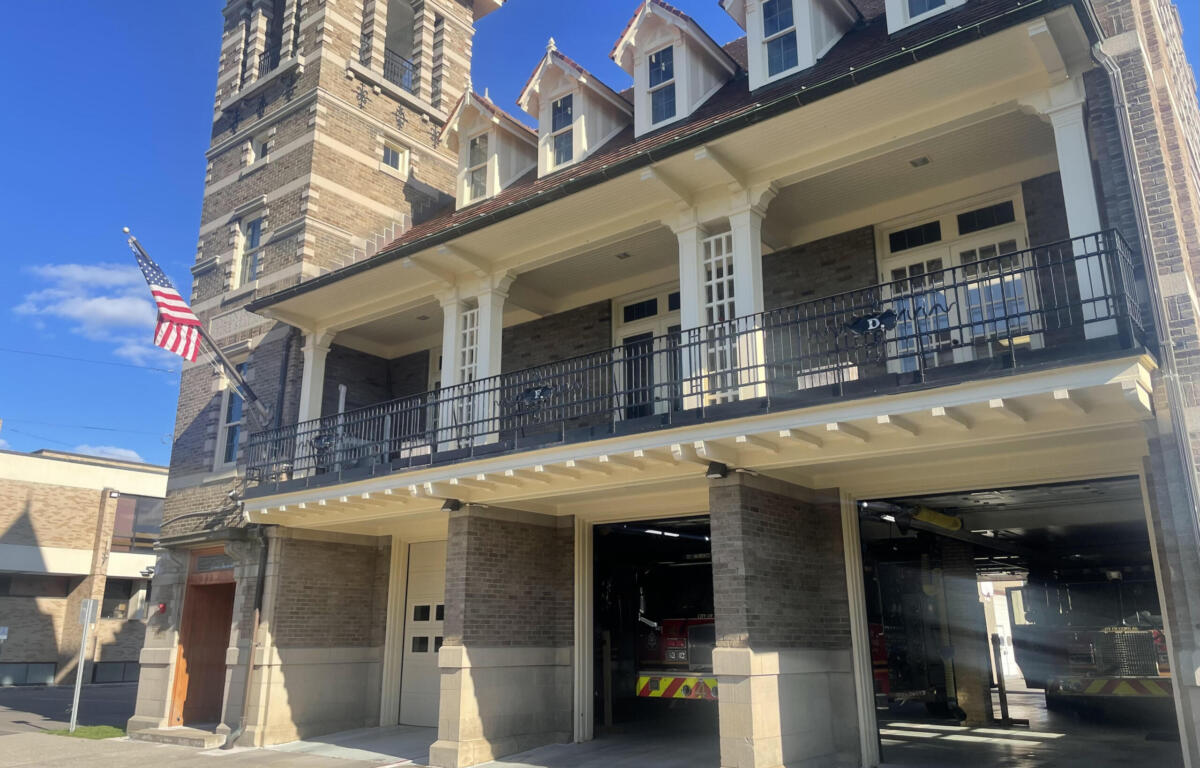 Brick fire station exterior with white columns, black balcony railing, and an American flag; a fire engine is visible in the open garage bay.