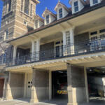 Brick fire station exterior with white columns, black balcony railing, and an American flag; a fire engine is visible in the open garage bay.