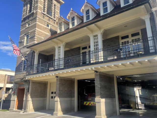 Brick fire station exterior with white columns, black balcony railing, and an American flag; a fire engine is visible in the open garage bay.