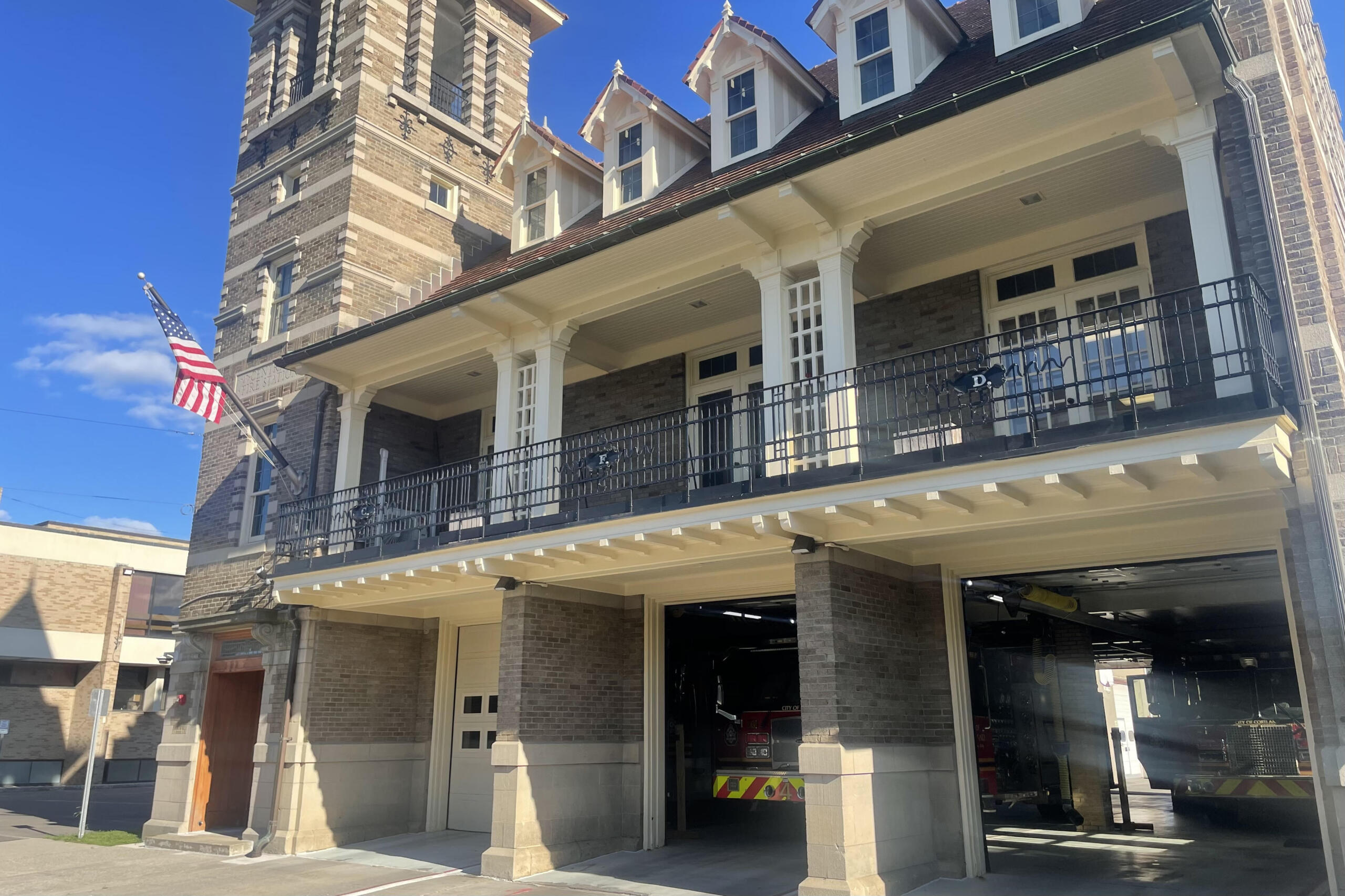 Brick fire station exterior with white columns, black balcony railing, and an American flag; a fire engine is visible in the open garage bay.